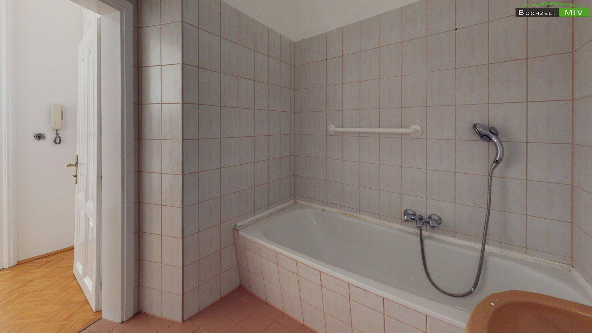 Badezimmer mit Badewanne und hellen Fliesen, ausgestattet mit einer Duscharmatur und Haltegriff.