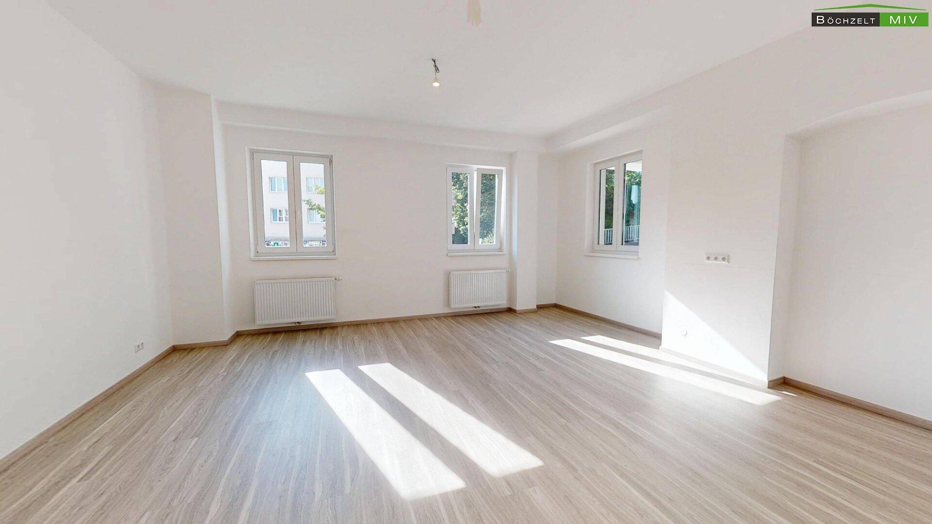 Helles, leeres Zimmer mit Laminatboden, weißen Wänden und mehreren Fenstern, durch die Sonnenlicht fällt.