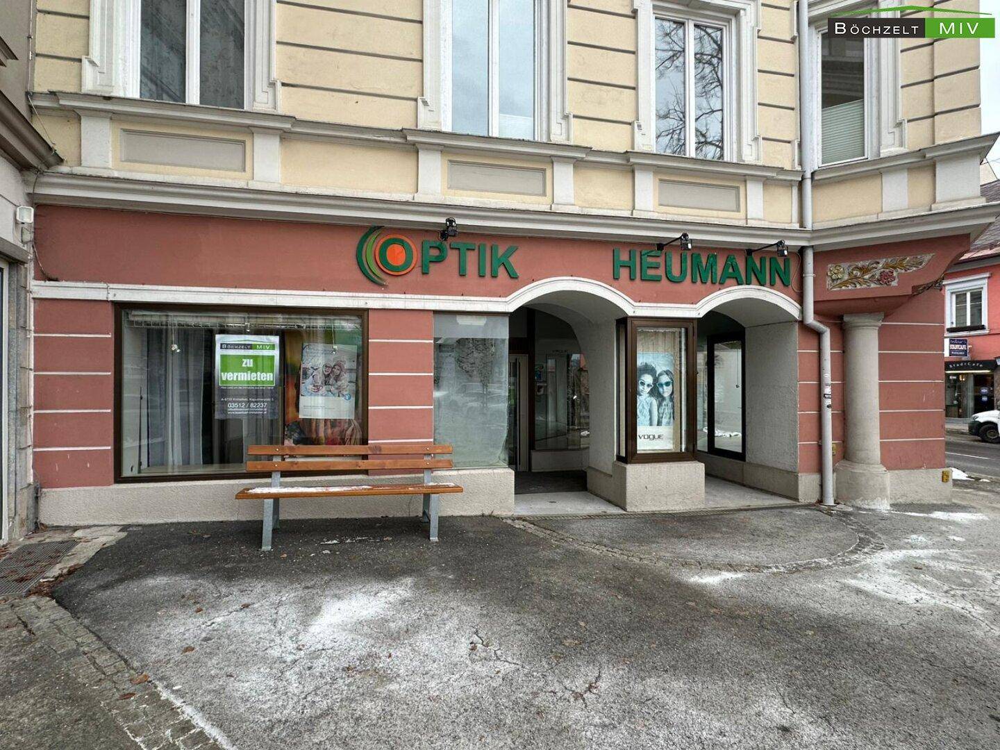 Außenansicht eines Geschäftsgebäudes mit traditioneller Fassade, großen Schaufenstern und einer Sitzbank davor.