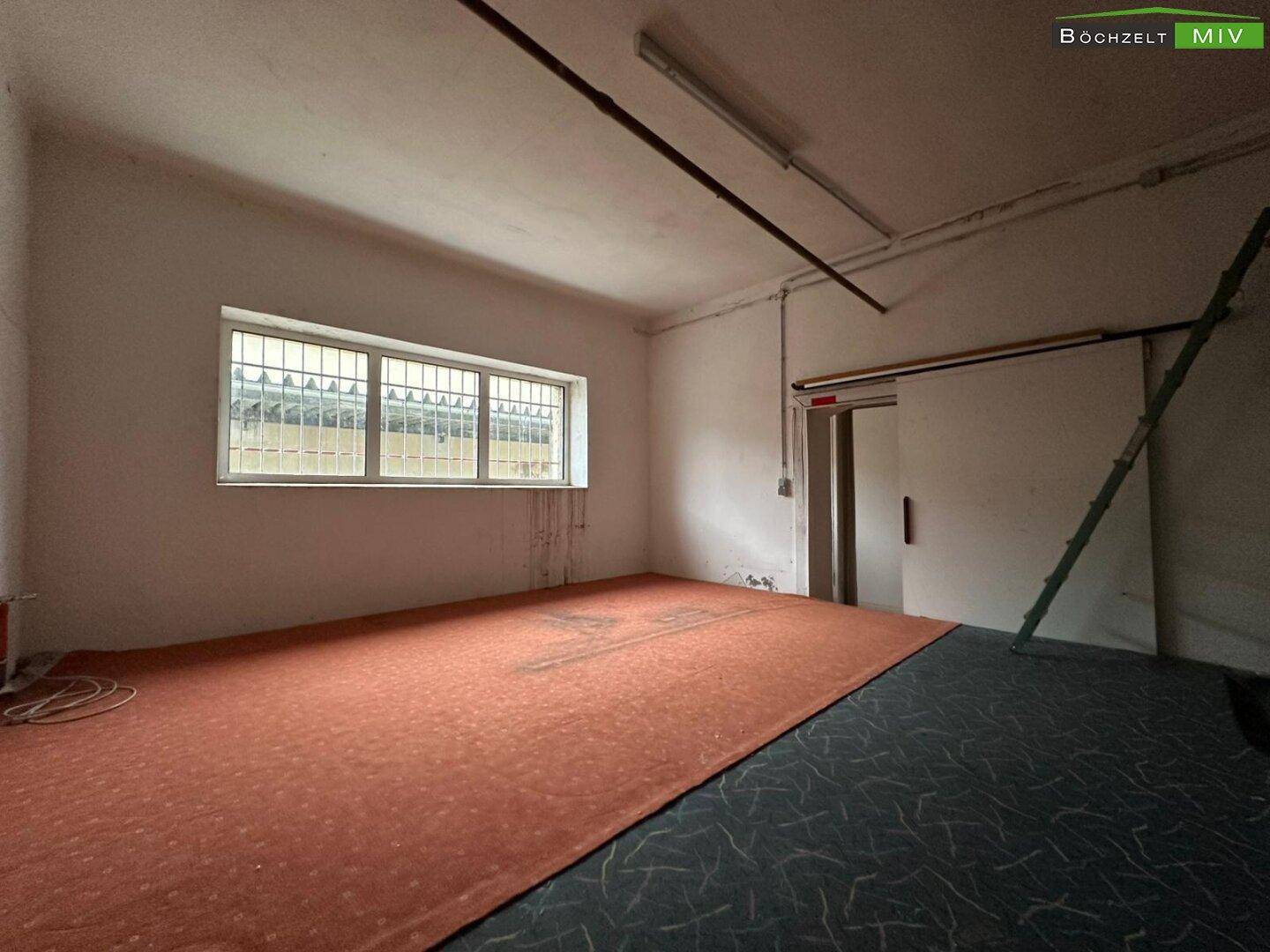 Innenraum eines Gewerbeobjekts mit Teppichboden, einem Fenster mit Gitter und einfacher Beleuchtung.