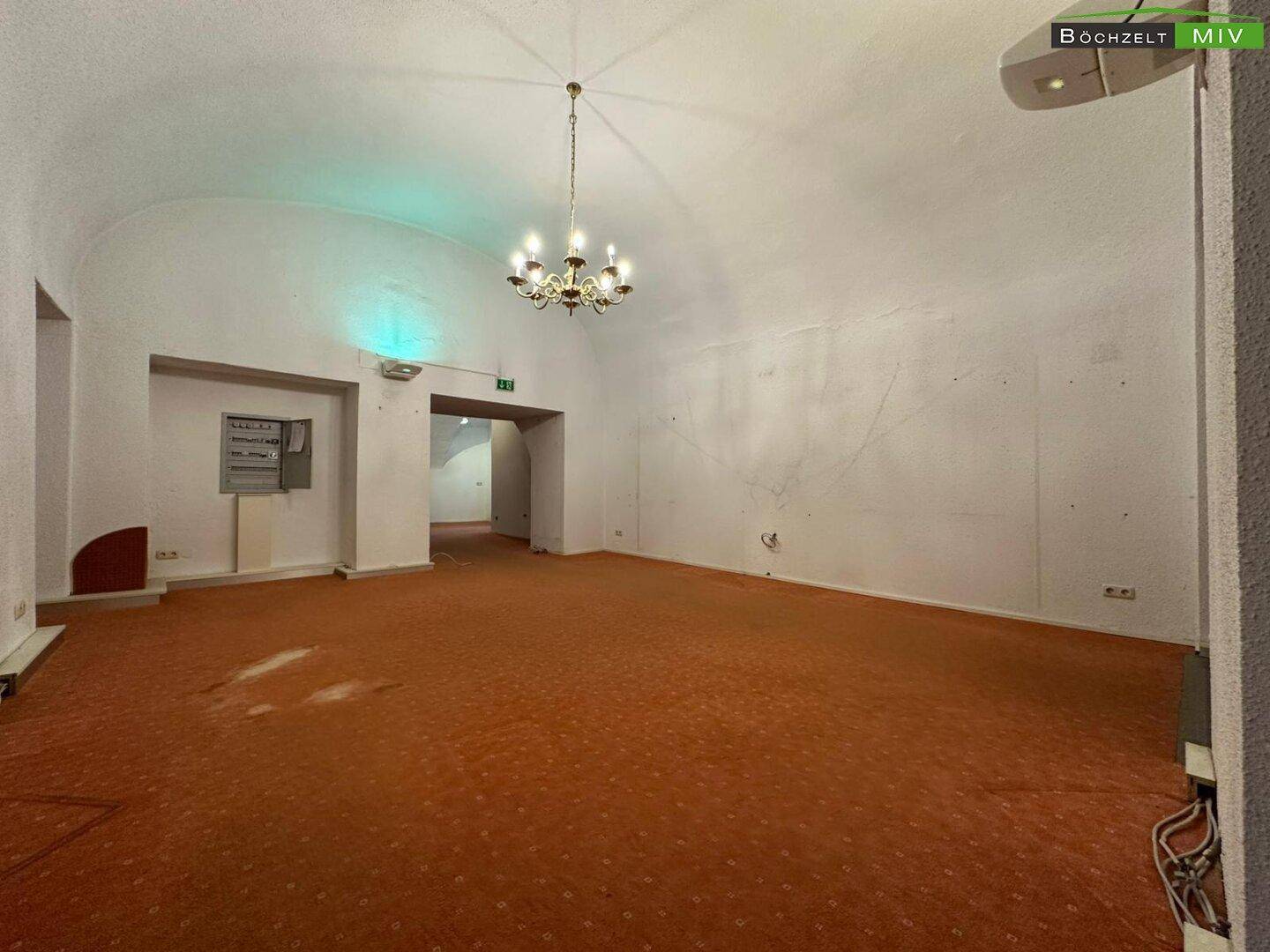 Großer, offener Gewerberaum mit Teppichboden, gewölbter Decke und einem Kronleuchter als Beleuchtung.