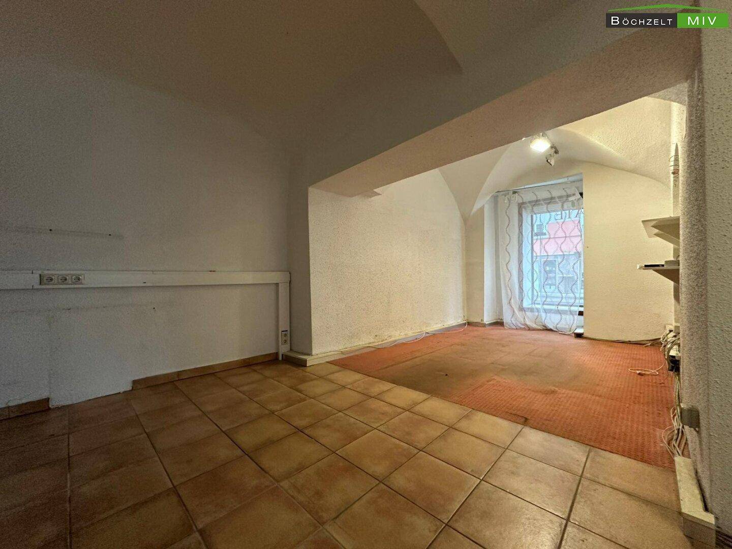 Hinterer Bereich eines Gewerbeobjekts mit Fliesen- und Teppichboden, einem Fenster und einfacher Beleuchtung.