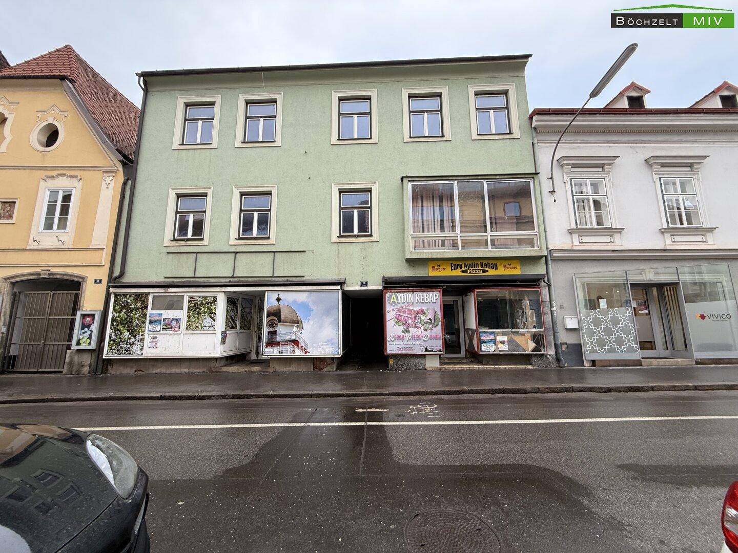 Ansicht des grünen Gebäudes mit Ladenlokalen im Erdgeschoss und Fenstern der oberen Stockwerke.