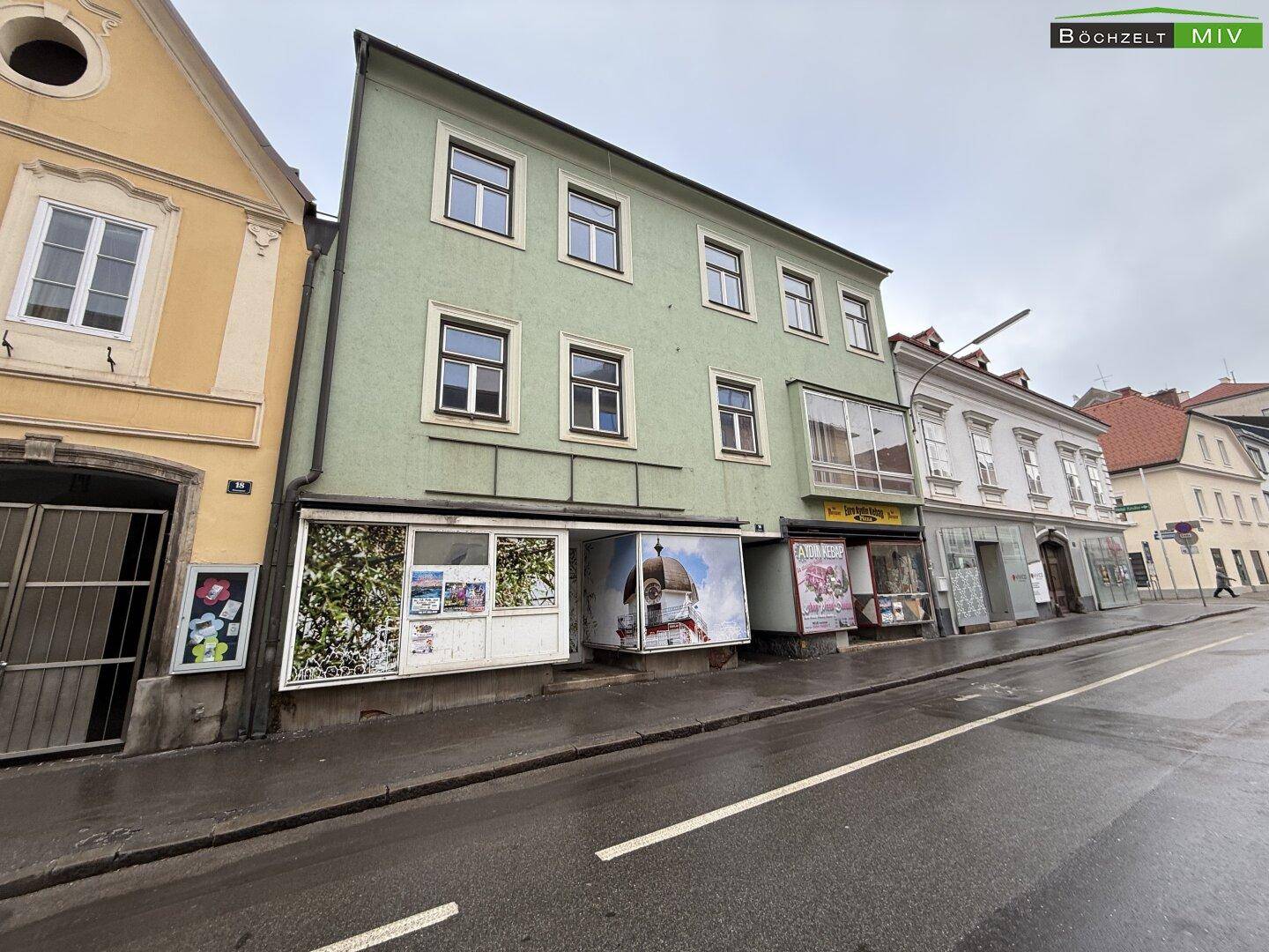 Grünes Gebäude mit Geschäften im Erdgeschoss und Wohnungen in den oberen Etagen, an einer belebten Straße gelegen.