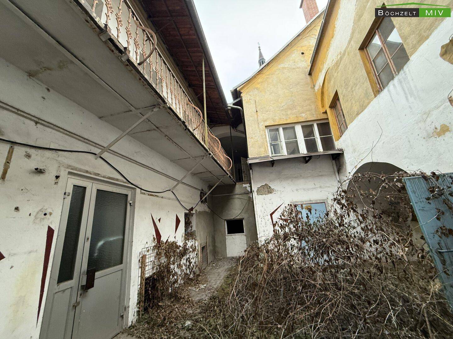 Alter Innenhof mit verfallenen Gebäudefassaden, überwucherten Pflanzen und einem Balkon im Obergeschoss.