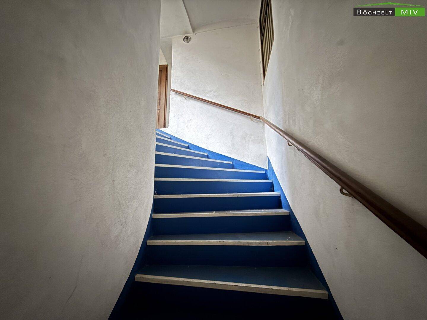 Enges Treppenhaus mit blauen Stufen und einem Holzgeländer, das in die oberen Stockwerke führt.