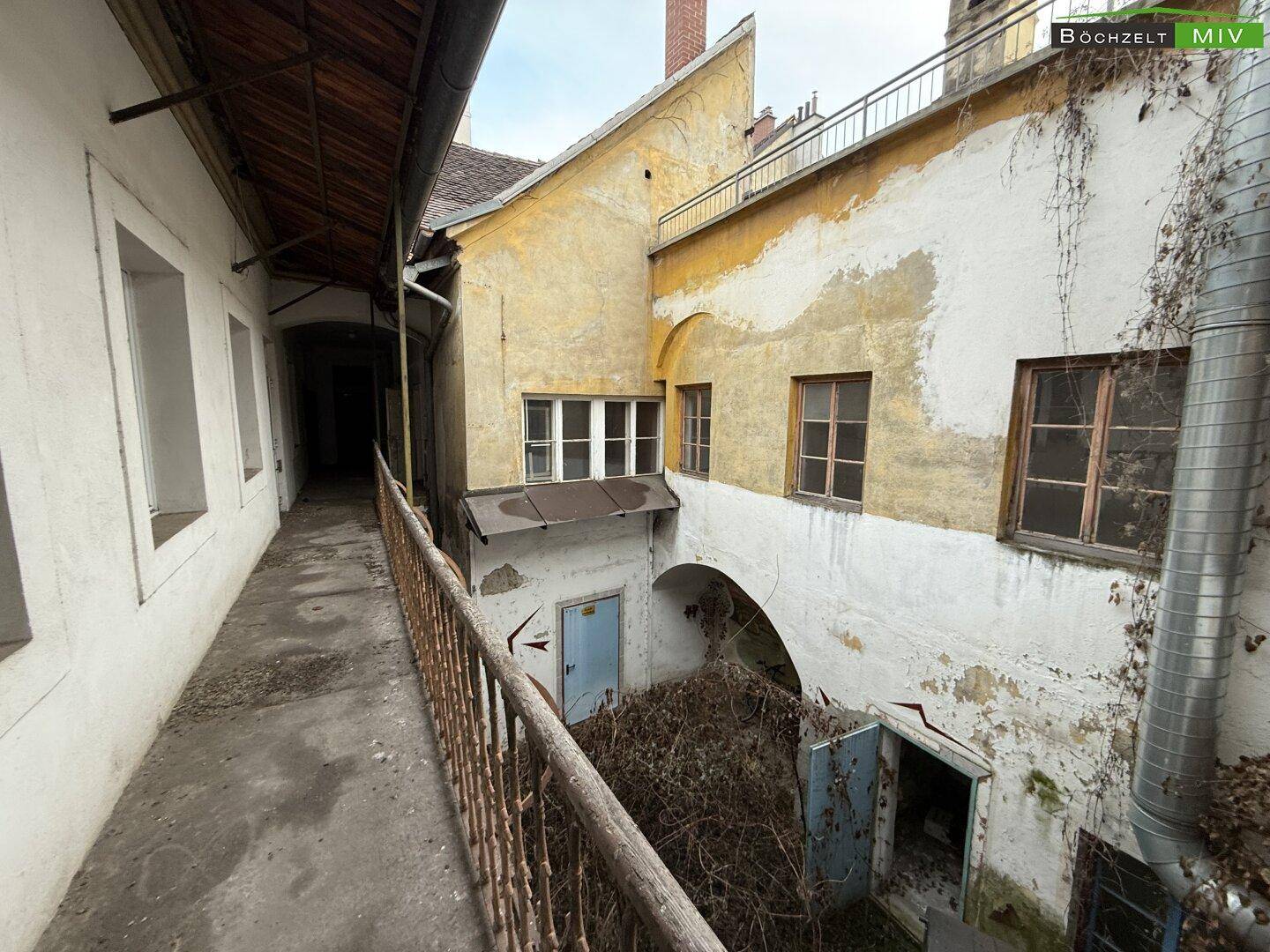 Blick auf einen verwilderten Innenhof mit alten Gebäuden, verrostetem Geländer und überwucherten Pflanzen.
