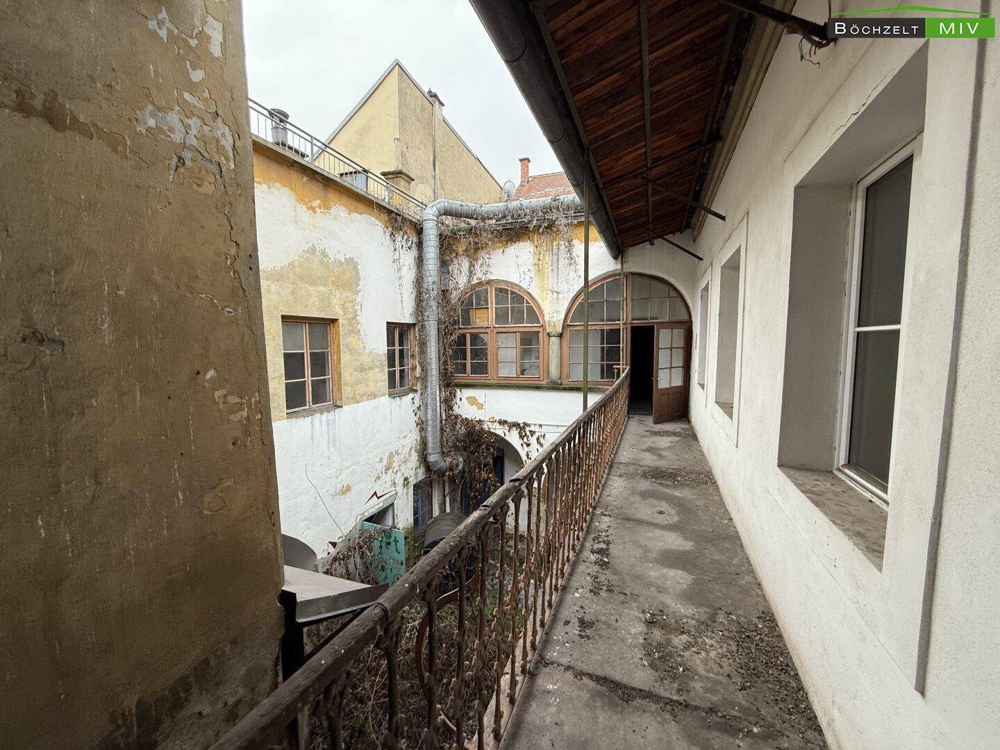 Überdachter Gang mit Blick auf einen Innenhof, gekennzeichnet durch alte Fenster und verrostete Geländer.
