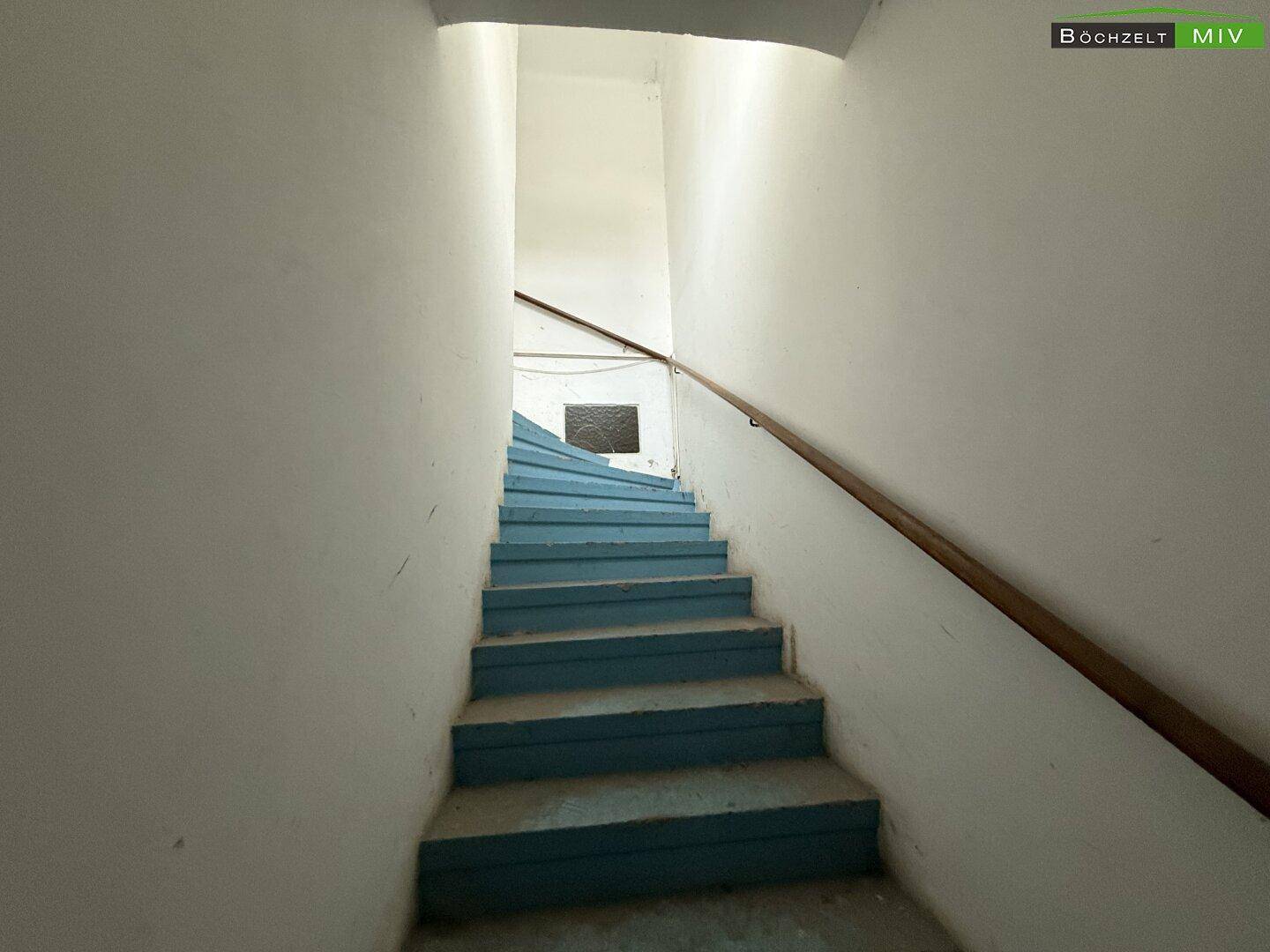 Enges Treppenhaus mit blauen Stufen und einem Holzgeländer, das in die oberen Stockwerke führt.