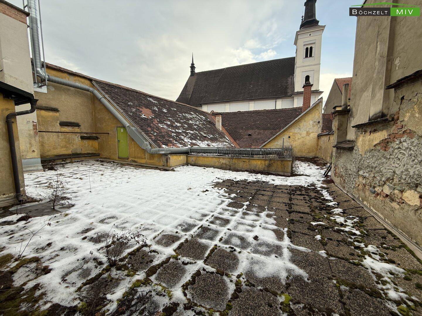 Große gepflasterte Terrasse mit Schnee und Blick auf eine historische Kirche im Hintergrund.