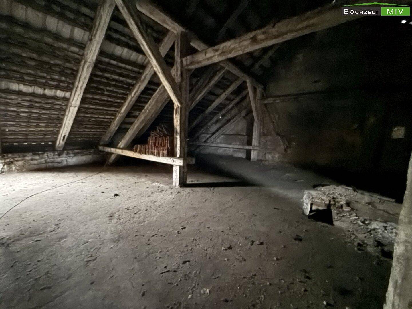 Dunkler Dachboden mit massiven Holzbalken und unfertigem Boden, der auf Ausbau wartet.