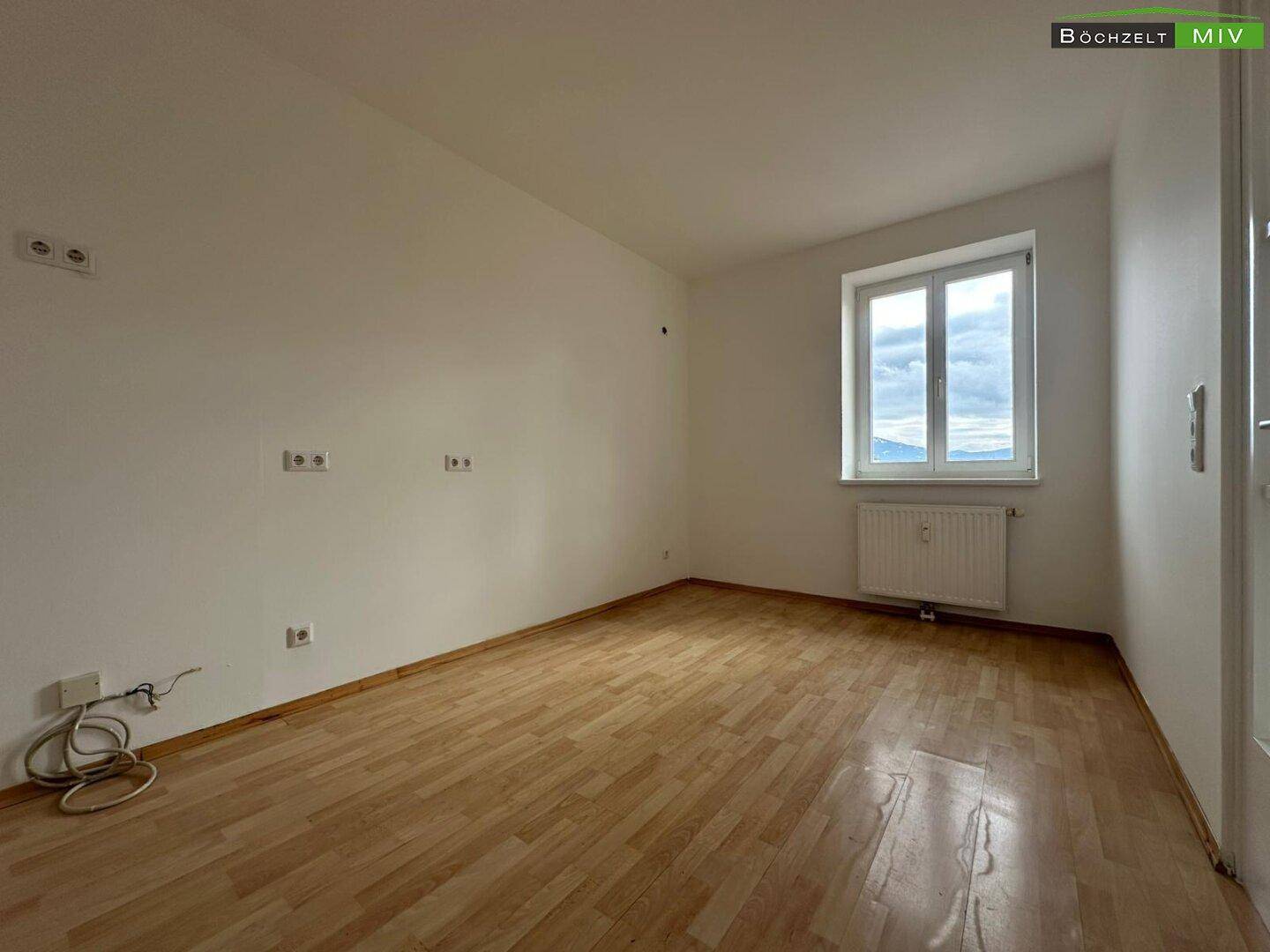Leeres Schlafzimmer mit hellem Parkettboden, weißen Wänden und Fenster mit Bergblick.