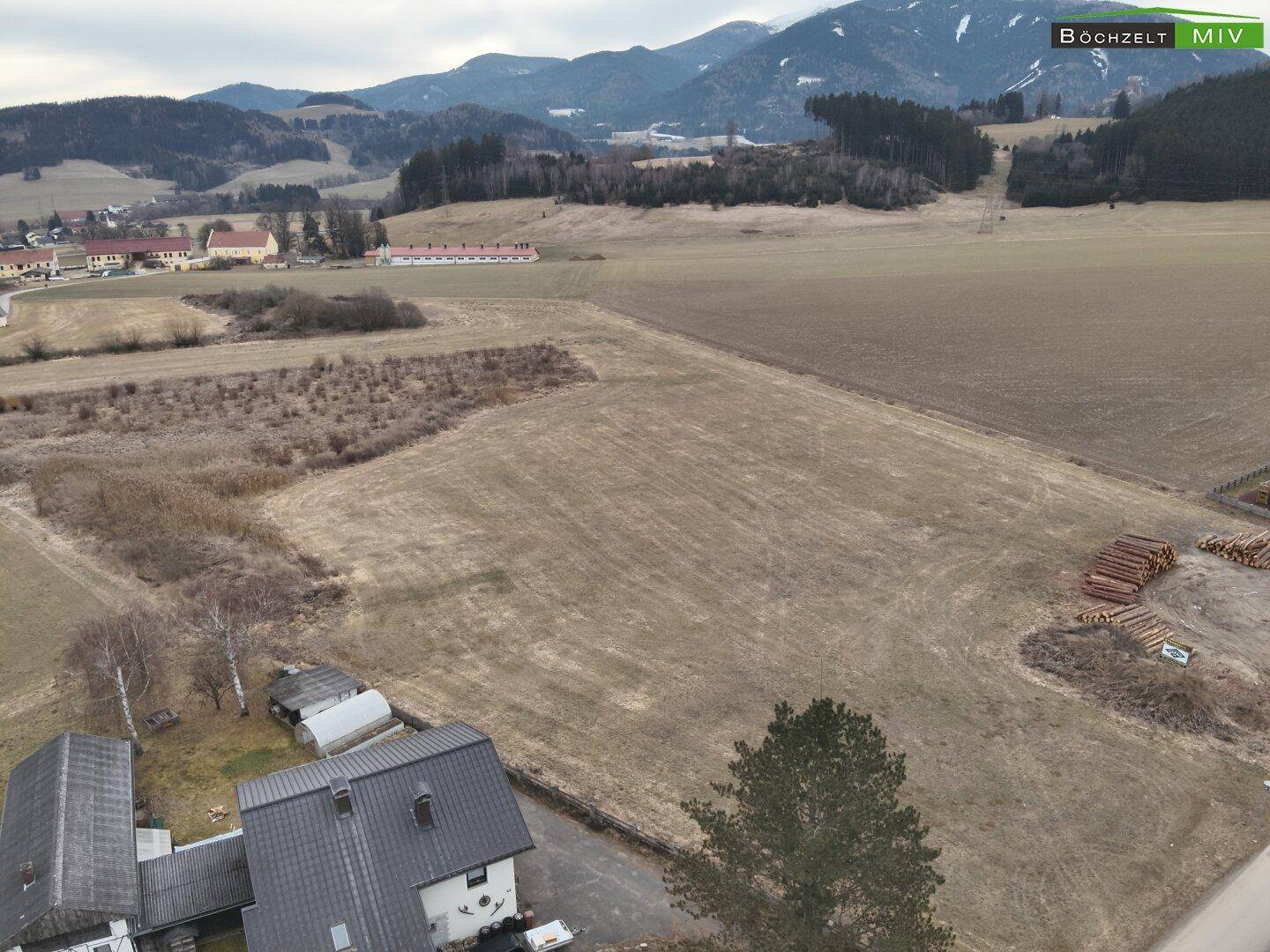 Weitläufige Landschaft mit Feldern, Häusern und Bergen im Hintergrund unter bewölktem Himmel.