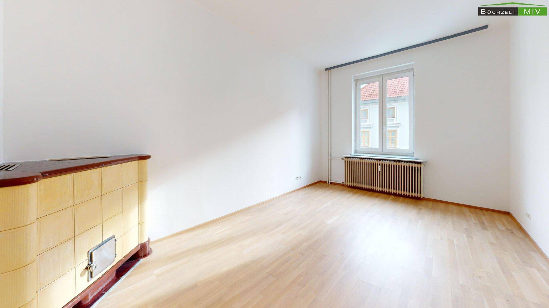 Helles Zimmer mit Parkettboden, Fenster und einem traditionellen Kachelofen.