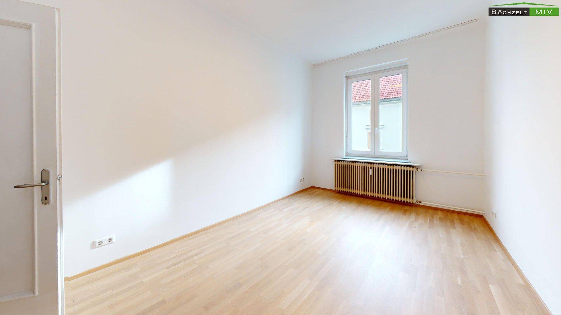 Lichtdurchflutetes Zimmer mit Parkettboden, weiß gestrichenen Wänden und Fenster.