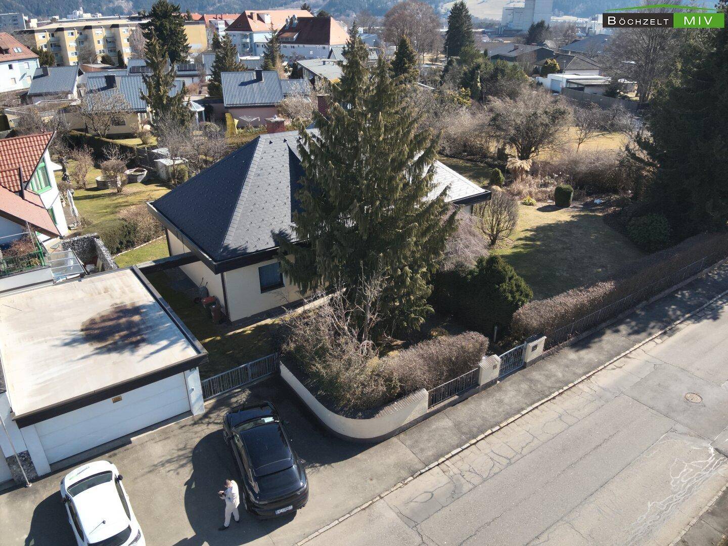 Luftaufnahme eines Hauses mit angrenzender Garage und Zufahrt, umgeben von Bäumen und Nachbarhäusern.