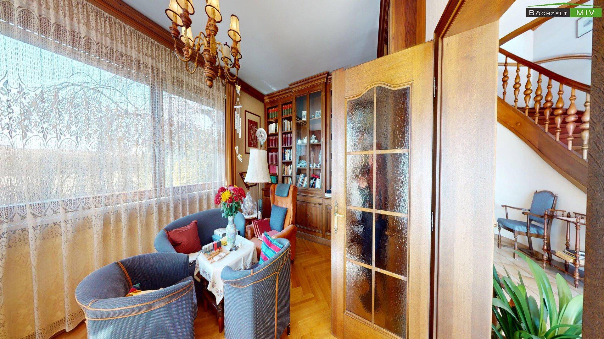 Gemütliches Wohnzimmer mit Parkettboden, Bücherregal und Blick in den Garten durch große Fenster.