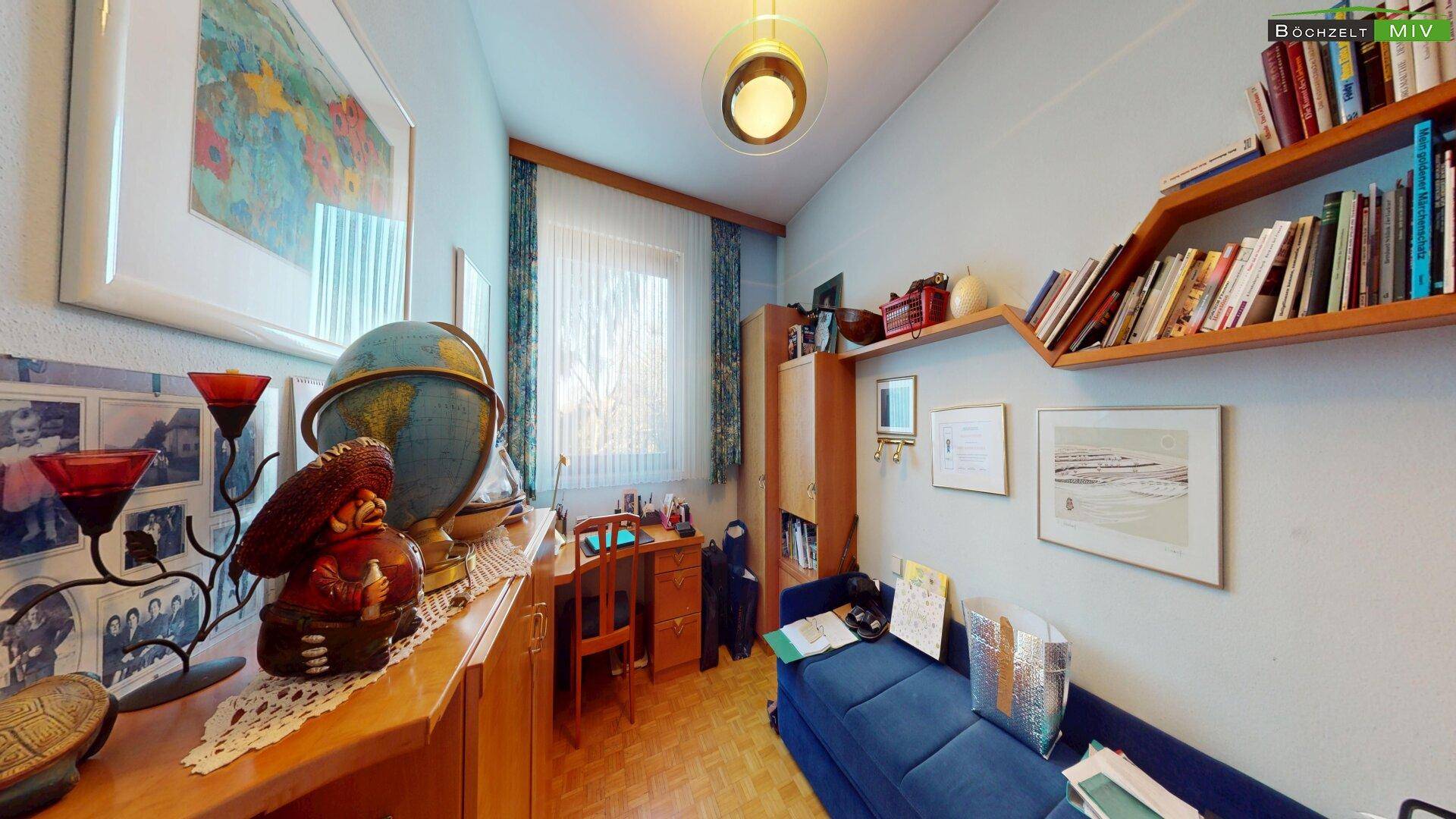 Arbeitszimmer mit Schreibtisch, Bücherregalen und einem blauen Sofa, gut beleuchtet.