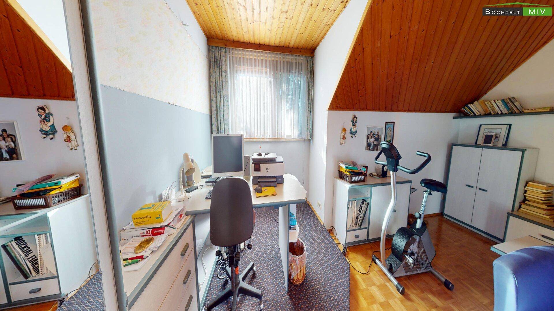 Arbeitszimmer im Dachgeschoss mit Schreibtisch, Sportgerät und Holzdecke.