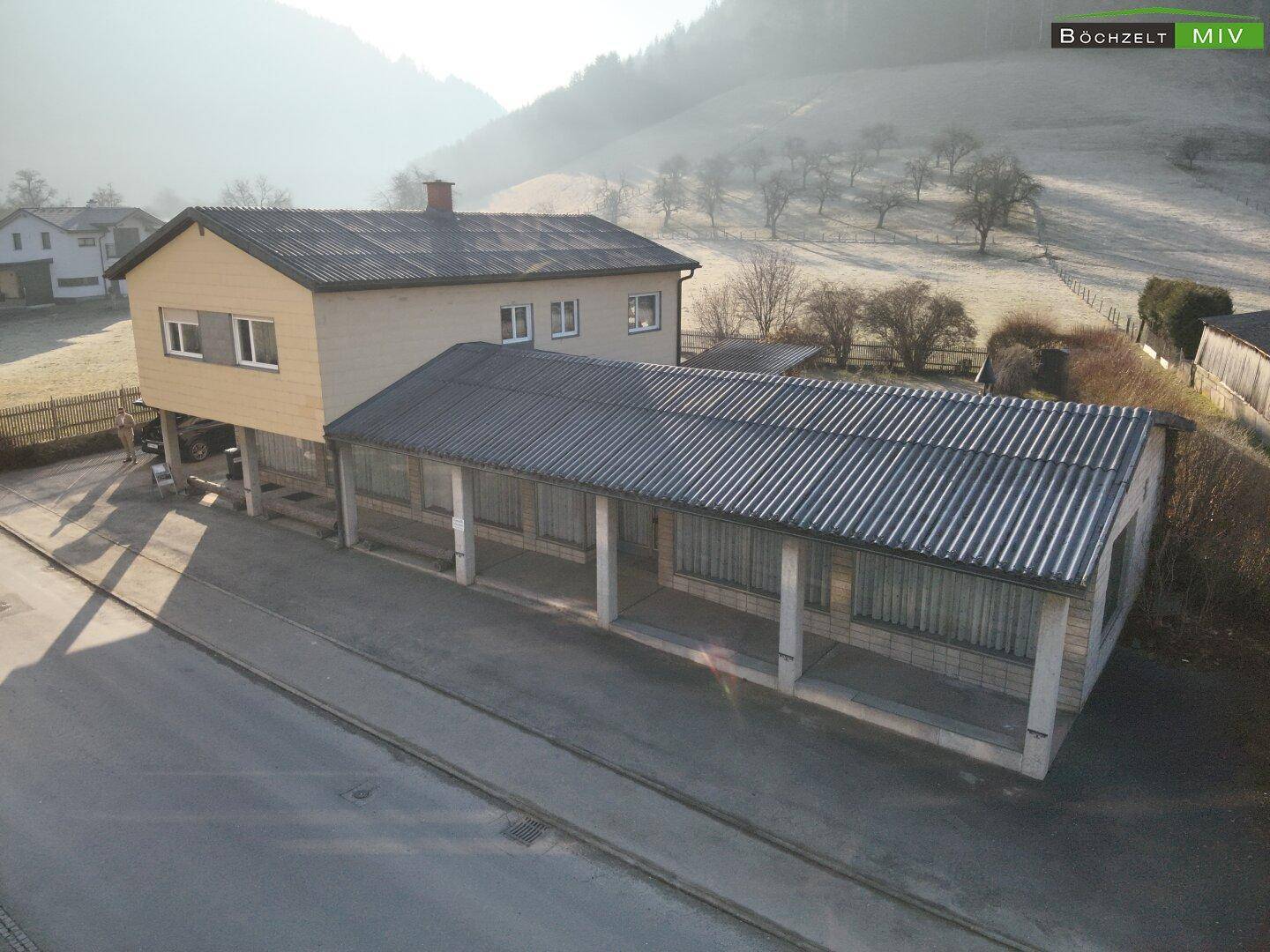 Ansicht des Hauses mit einem langen Anbau und der Straße im Vordergrund, umgeben von Natur.