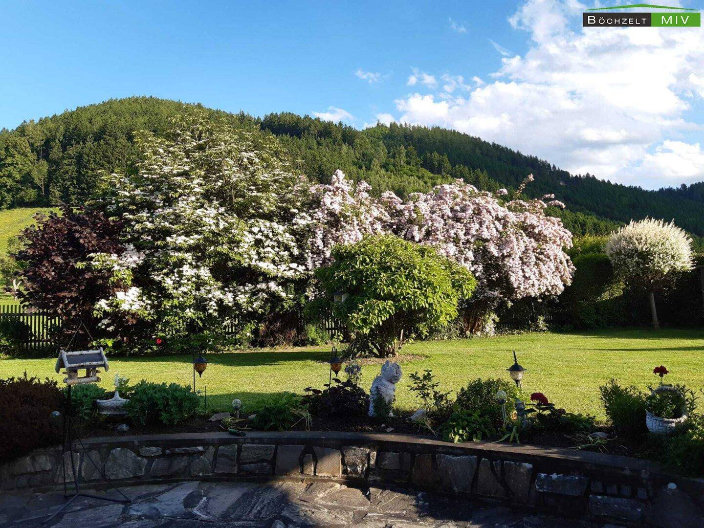 Gepflegter Garten mit üppiger Vegetation, blühenden Sträuchern und einem weiten Blick auf die Hügel.