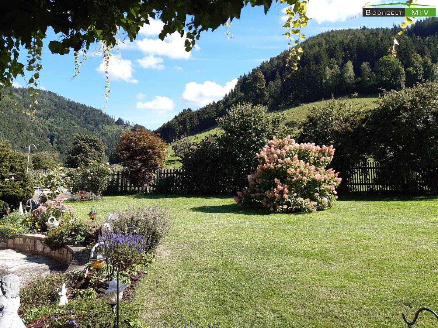 Sonniger Garten mit Rasenfläche, blühenden Sträuchern und einem weiten Blick auf die Landschaft.