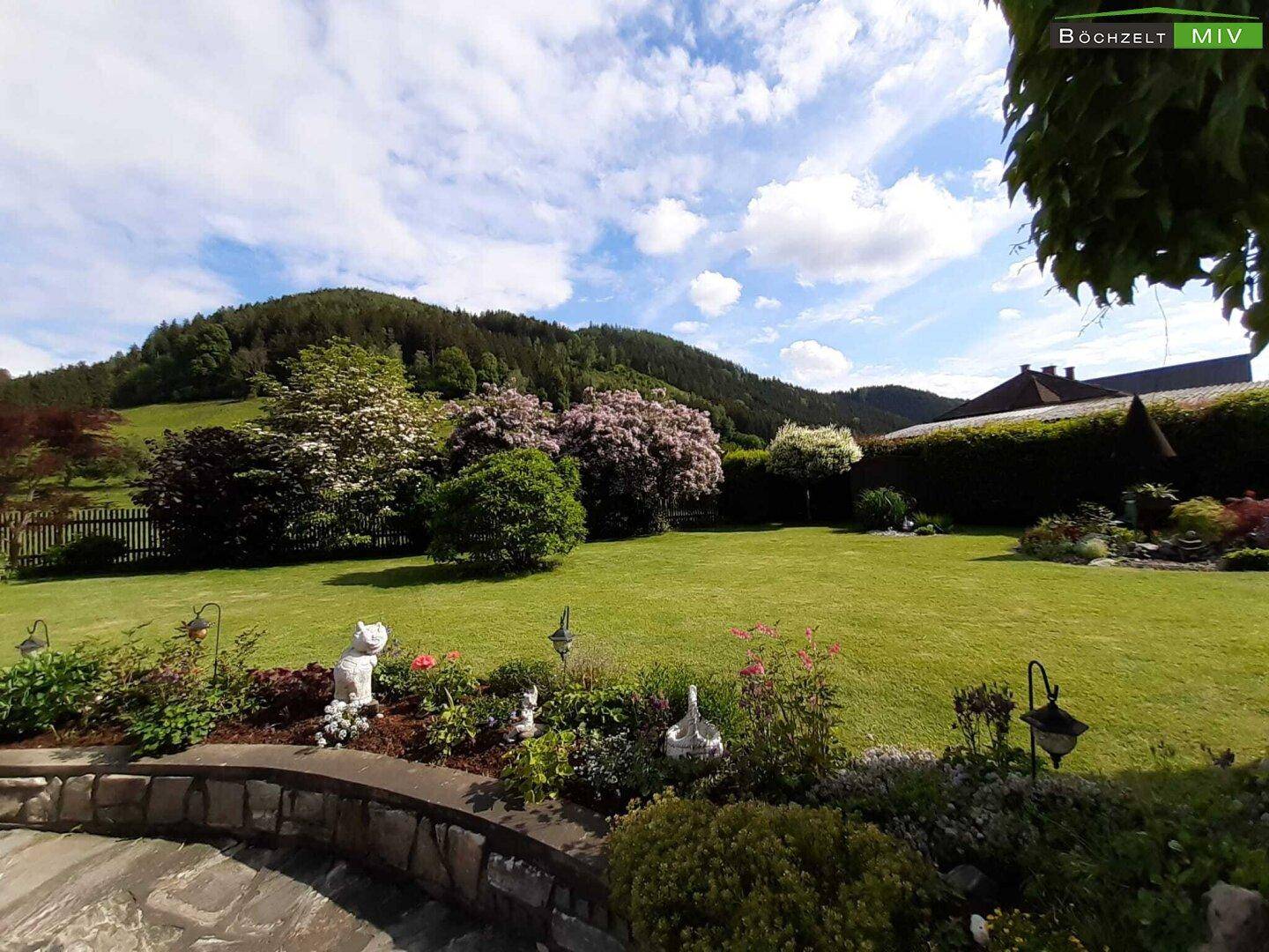 Weitläufiger Garten mit Rasen, Blumenbeeten und Blick auf die bewaldeten Hügel unter blauem Himmel.