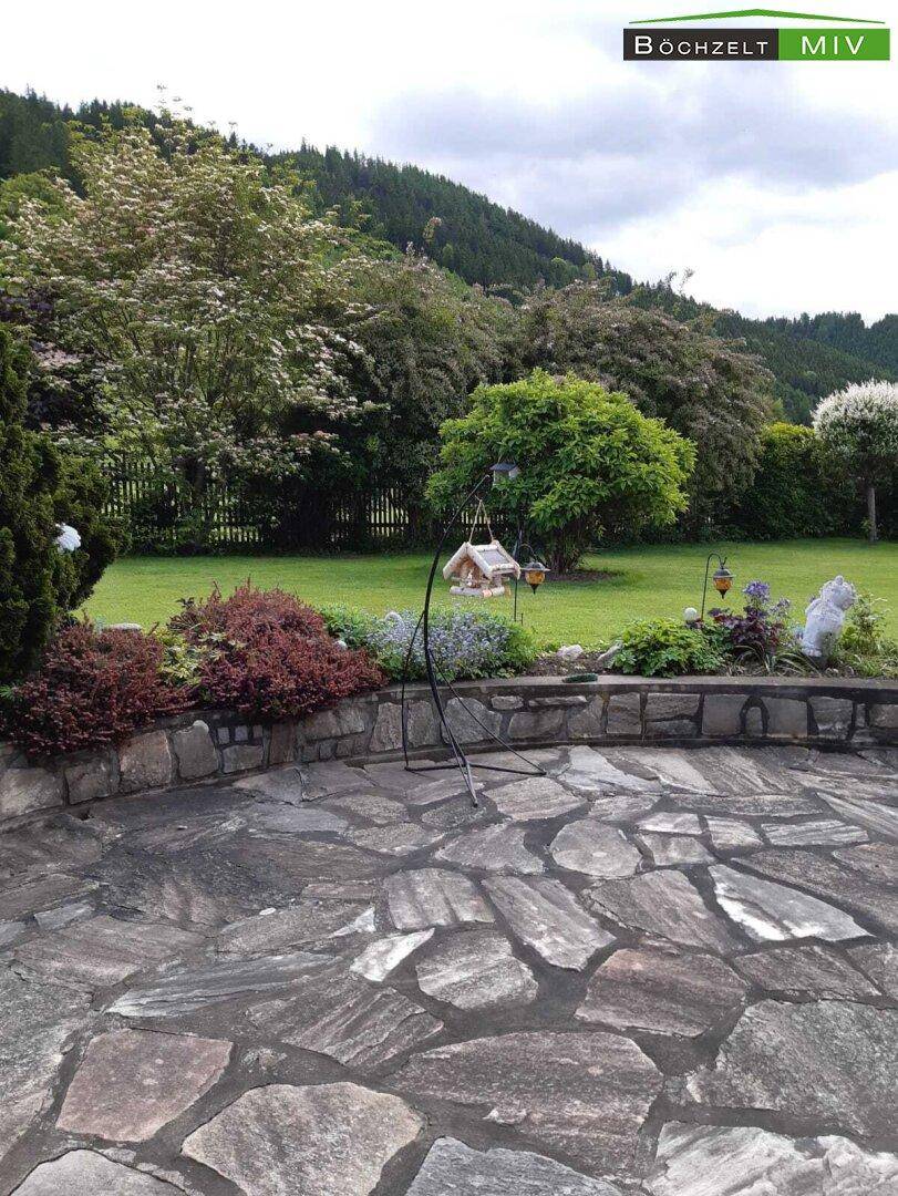 Gepflegter Garten mit Steinterrasse, Rasenfläche und Blick auf die umliegende Natur.