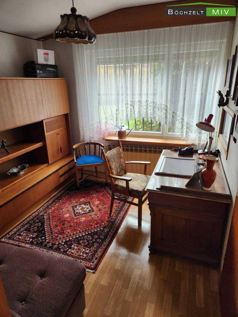 Arbeitszimmer mit Holzschreibtisch, Bürostuhl und einem Teppich, hell durch großes Fenster.