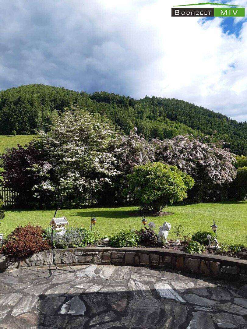 Weitläufiger Garten mit Rasen, blühenden Sträuchern und Blick auf die bewaldeten Hügel.