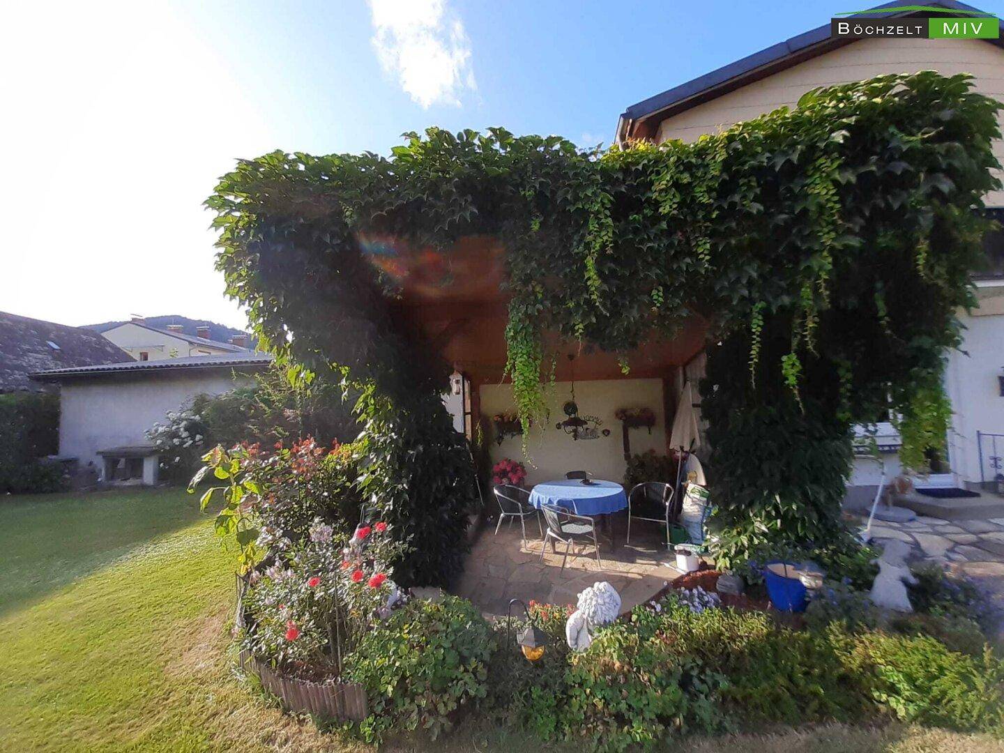 Überdachte Terrasse mit Sitzgelegenheiten, umgeben von üppigem Grün und Blumen.