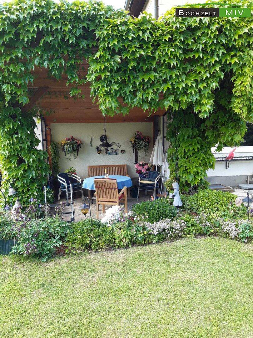 Gemütliche überdachte Terrasse mit Gartenmöbeln, umrankt von Kletterpflanzen und Blumen.