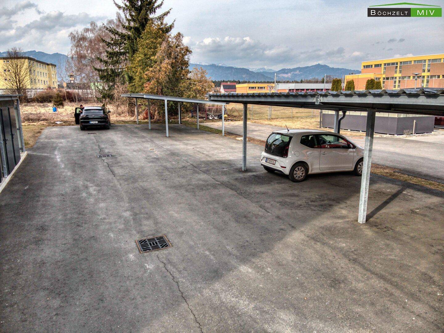 Außenparkplätze mit Carports vor einem Wohngebäude in ländlicher Umgebung.