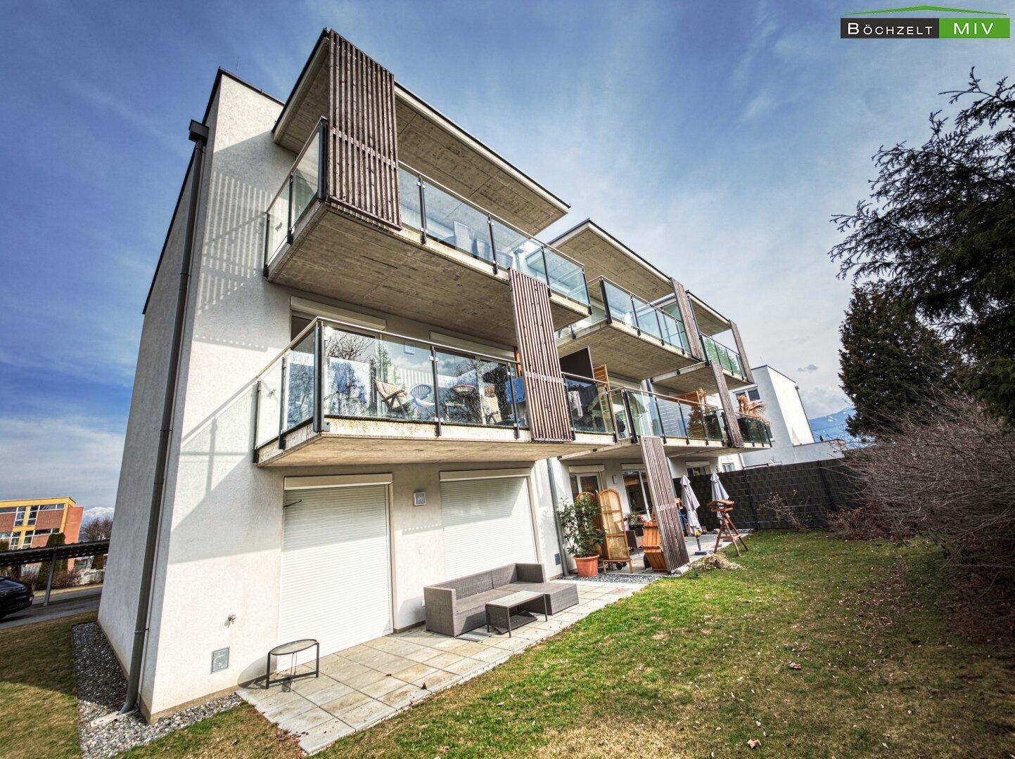 Seitenansicht eines modernen Wohngebäudes mit Balkonen und einer kleinen gepflasterten Terrasse.