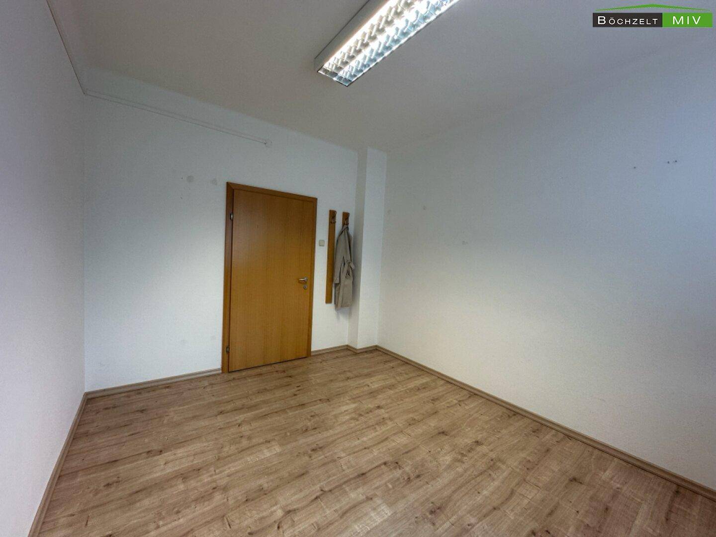 Leerer Büroraum mit Laminatboden und einer Holztür, bereit zur individuellen Gestaltung und Nutzung.