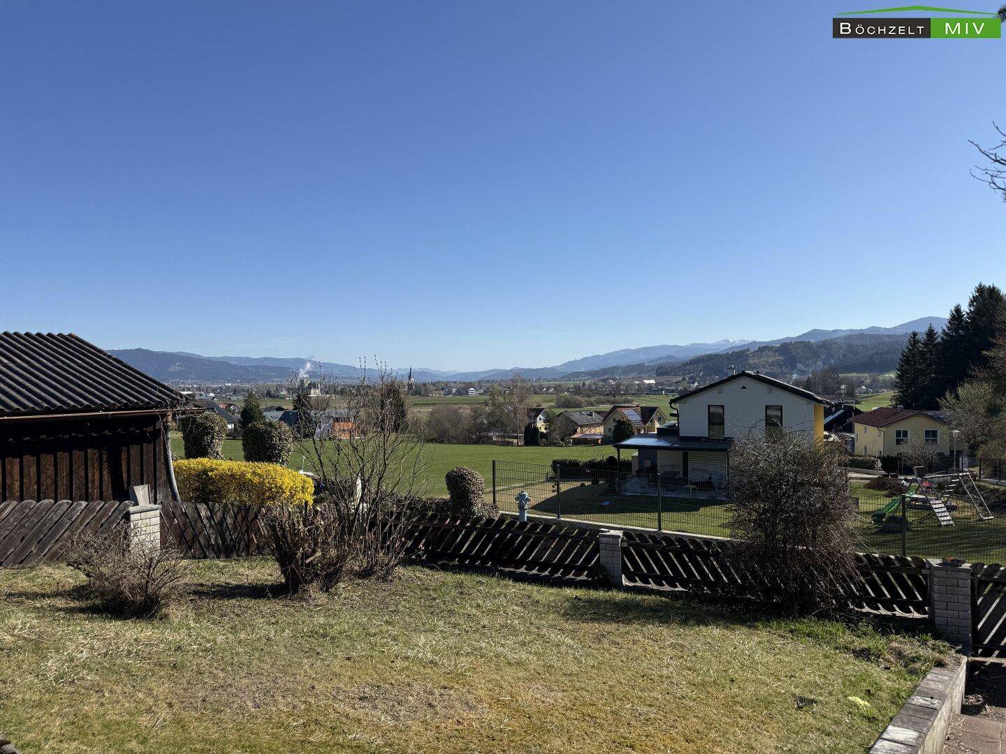 Sonniger Gartenbereich mit Rasenfläche, Holzzaun und weitem Blick über die ländliche Umgebung und Berge.