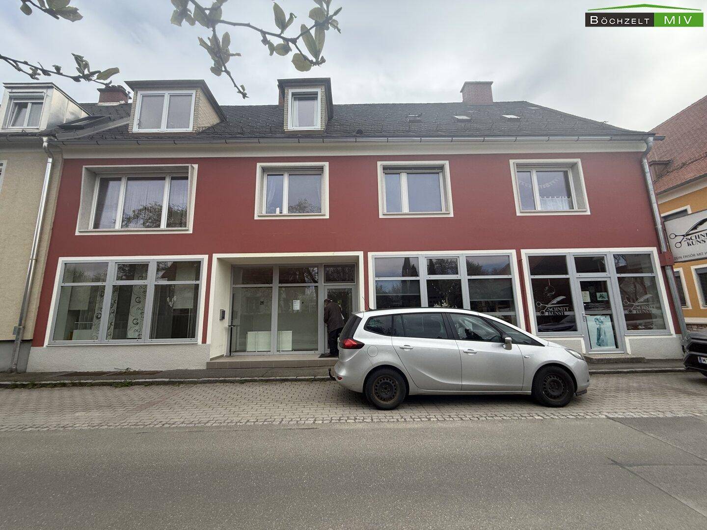 Ansicht der roten Gebäudefassade mit großen Fenstern und einem silbernen Auto auf der Straße geparkt.