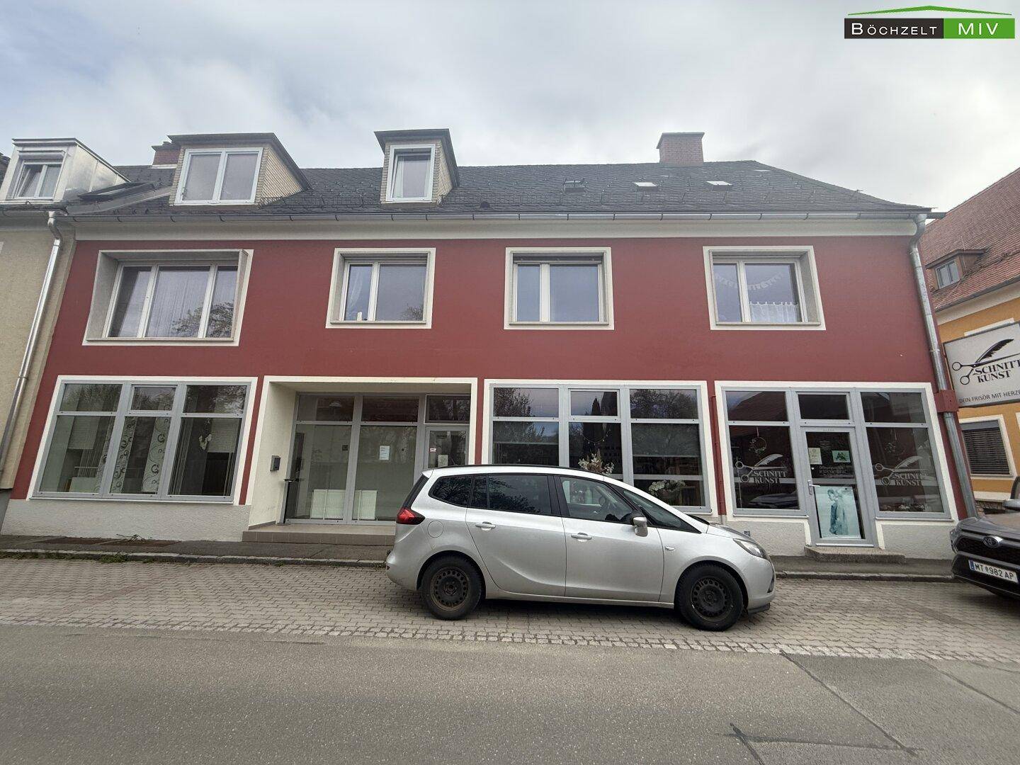 Frontansicht des roten Gebäudes mit großen Schaufenstern im Erdgeschoss und einem Auto davor.