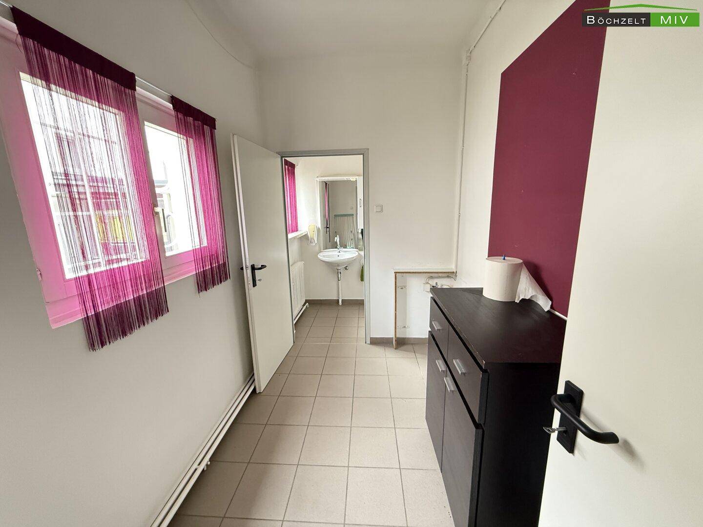 Raum mit Fliesenboden, Fenster mit Vorhängen und einem Schrank vor dem Badezimmer.