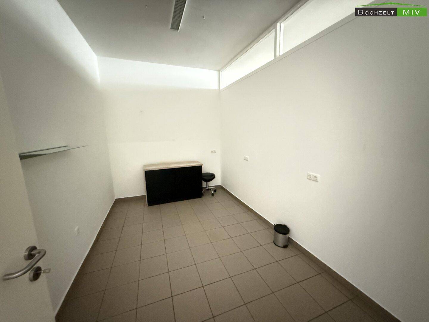 Leerer Büroraum mit Fliesenboden, weißen Wänden und einem kleinen Fensterband oben.