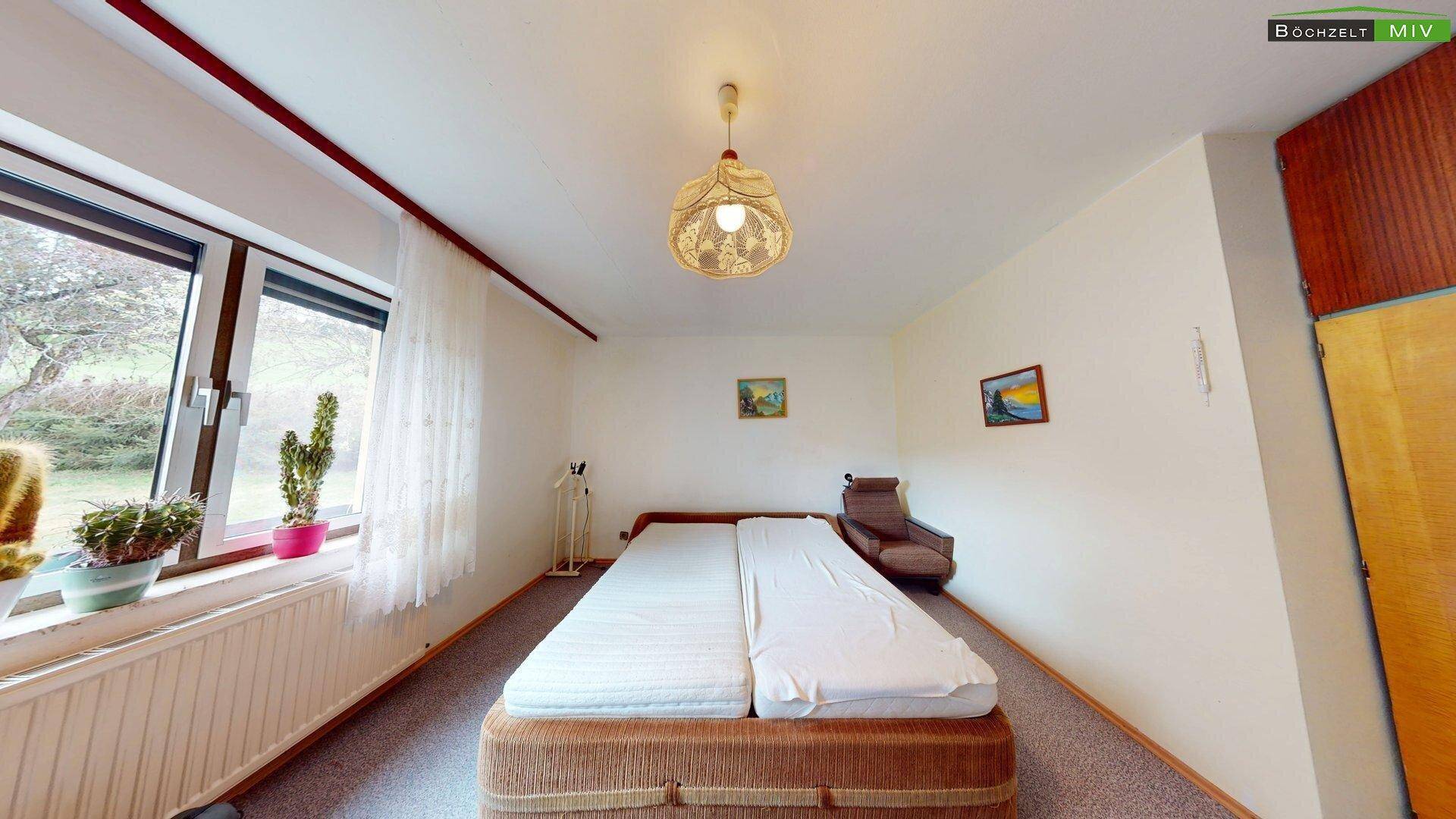 Helles Schlafzimmer mit Doppelbett, Teppichboden und Fenster mit Blick ins Grüne, einfach eingerichtet.