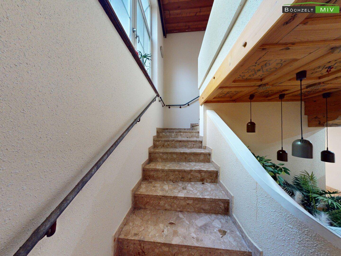 Steintreppe mit Metallgeländer führt nach oben, umgeben von hellen Wänden und Holzdecke.