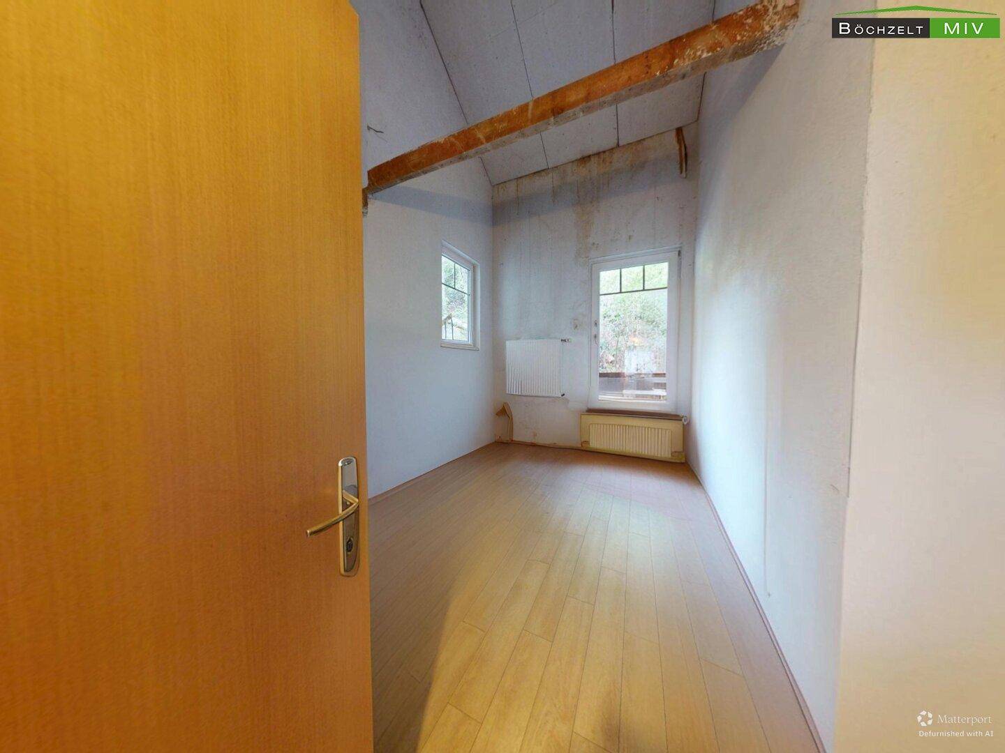 Leeres Zimmer mit Holzboden, sichtbarem Holzbalken und zwei Fenstern unter der Dachschräge.