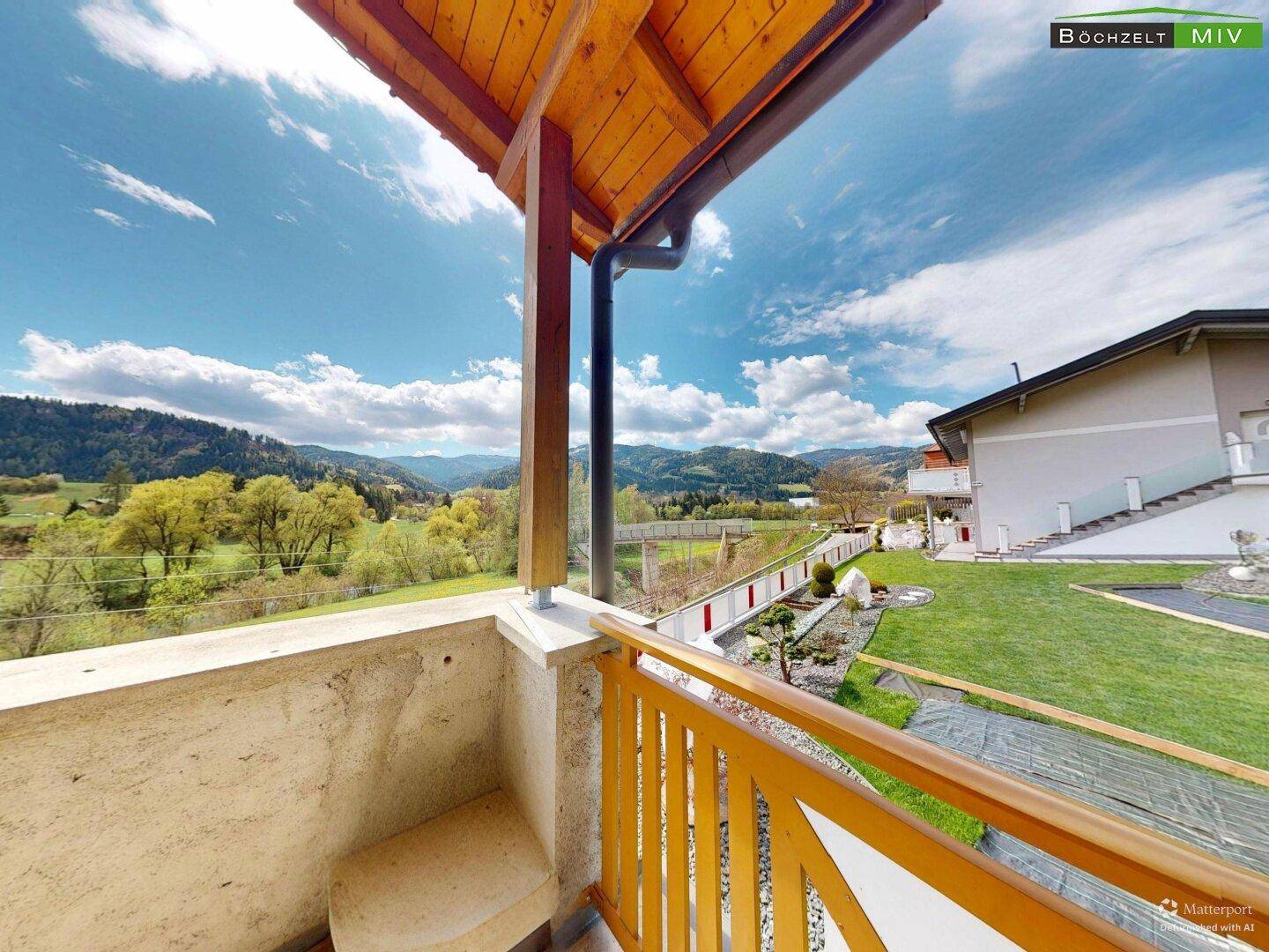 Überdachter Balkon mit Holzgeländer und weitem Blick auf die grüne Umgebung und Berge.