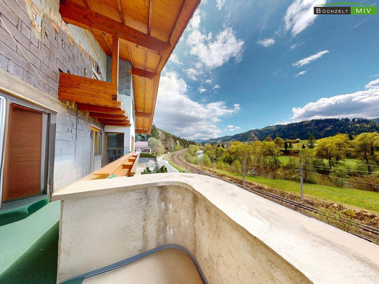 Langer Balkon mit Betonbrüstung und weitem Blick auf die Bahngleise und die Berglandschaft.