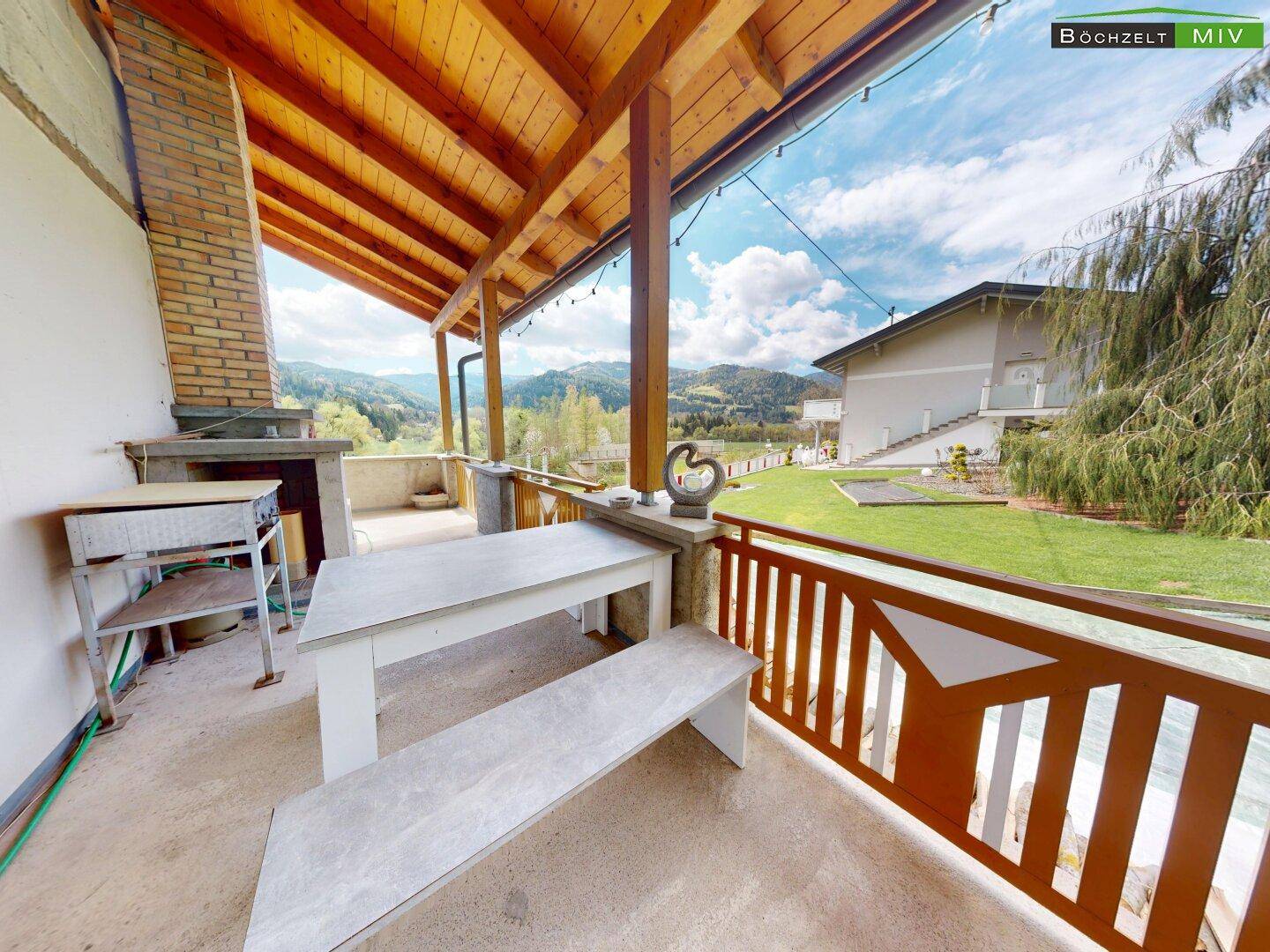 Überdachte Terrasse mit Sitzgelegenheiten und Grillplatz, umgeben von grüner Landschaft.