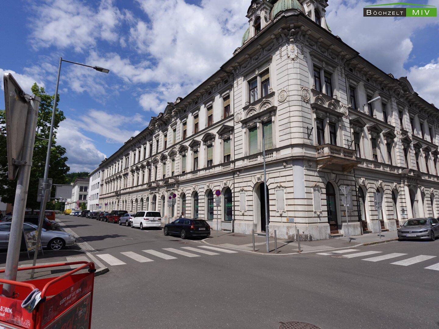 Historisches Eckgebäude mit detailreicher Fassade, umgeben von einer belebten Straße und geparkten Autos.