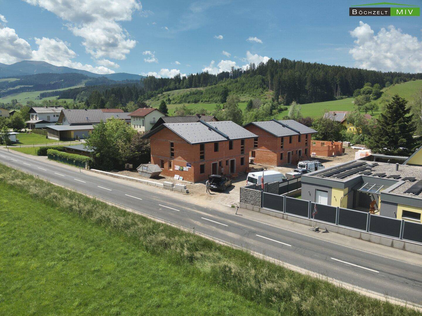 Luftaufnahme einer ländlichen Wohngegend mit Neubauten und grünen Hügeln unter blauem Himmel.