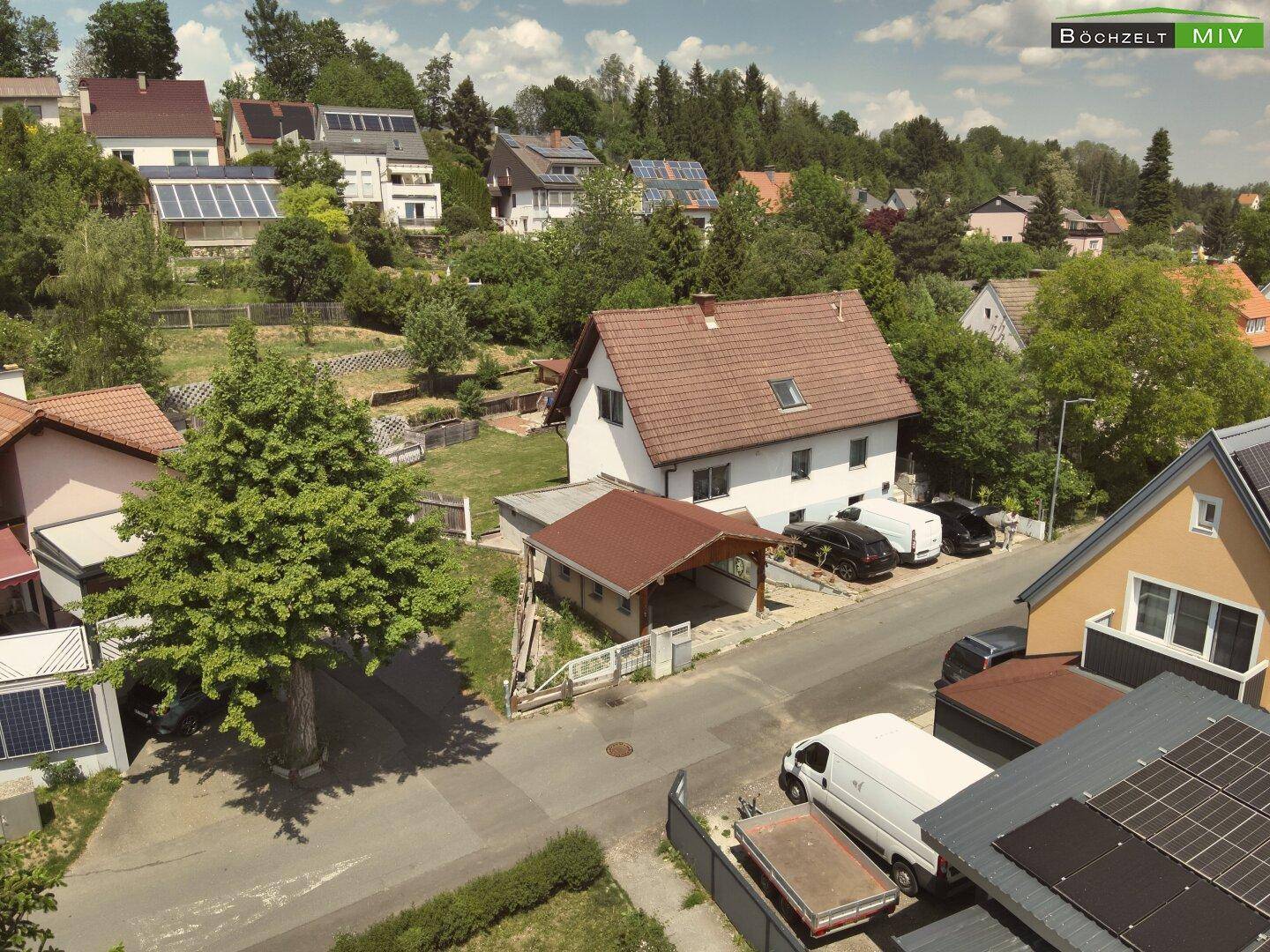 Panoramablick aus der Luft auf ein Wohnhaus mit Carport, umgeben von viel Grün und anderen Häusern.