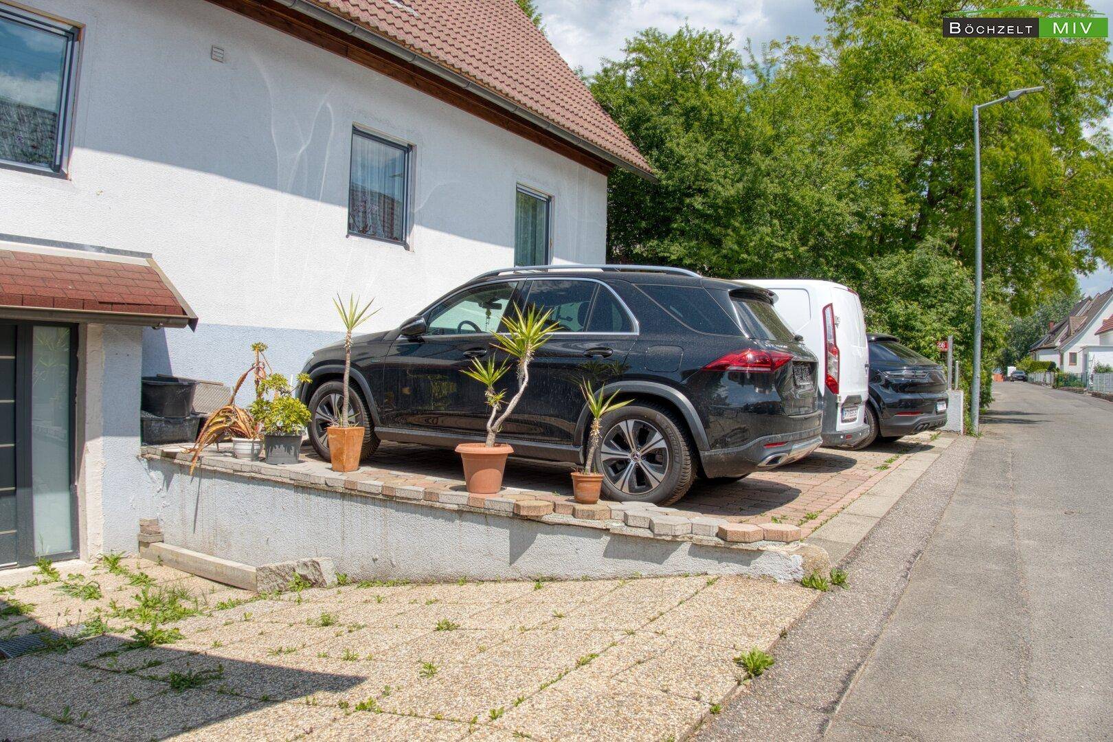 Vorderansicht des Hauses mit geparkten Fahrzeugen auf dem gepflasterten Stellplatz und Pflanzen.